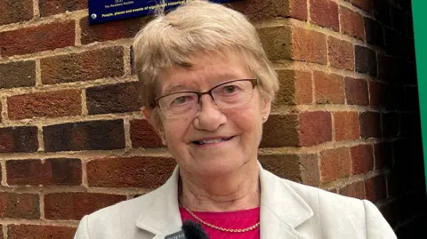 Sheila Welch has short, blonde hair and is wearing a white jacket and maroon top. She is also wearing glasses and is smiling at the camera.