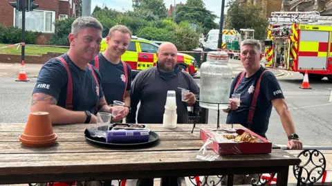 The Old Bank A group of four men from the fire service at a table outside a pub which has offered them refreshments. Behind them are fire service vehicles including a red and yellow fire engine and car.