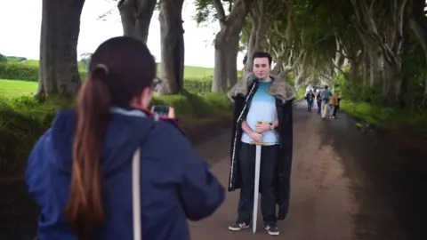 Getty Images A woman, facing away from the camera, taking a picture of a man wearing a cape and holding a sword. He is standing on a dirt path with large beech trees on either side.