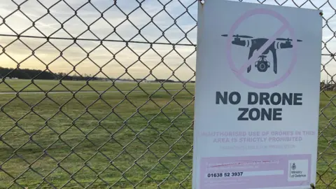 Matt Precey/BBC Sign prohibiting drones attached to the fence of RAF Lakenheath in Suffolk. Behind the fence is grass and a runway. The sign has an image of a drone.