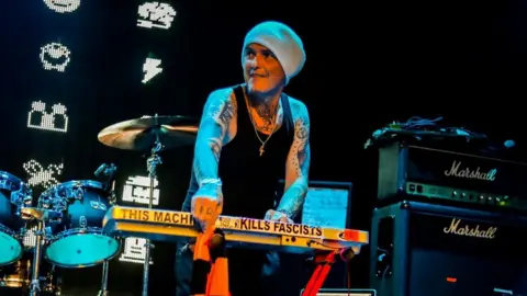 Derry Brownson, the keyboardist of EMF, smiles as he performs on stage. He is wearing a grey beanie, a black tank top and has arms covered in tattoos.