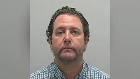 Bedfordshire Police Custody photo of Patterson wearing a blue, white and grey checked shirt. He is stood against a grey background, has slight stubble on his face and dark brown hair. 