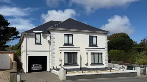 A white house, with a grey car in the garage. There is a road in front of it. The sky is blue with white clouds. 