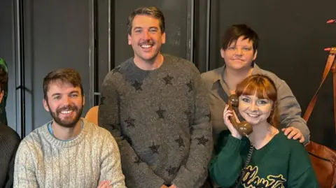 four people smiling at the camera in a posed shot; one is holding an old wired phone prop to her ear