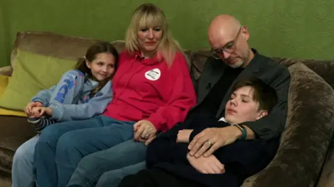 A family of four is sitting on a green sofa. Mother and father are sitting in the middle and their daughter is sitting on the left and their son is sitting on the right. 