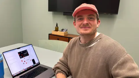 James smiles at the camera, next to an open laptop. He is wearing a beige jumper with a large hole in the collar and a pale pink cap. There is a screen on the wall in the background.