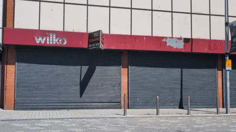 The Market Gallery The former Wilko store in South Shields. Large grey shutters cover the vacant retail units windows and doors. The red banner sign of the shop remains above the doors but it has been damaged and its paint has worn away in places. An estate agents sign has been installed advertising it for sale.