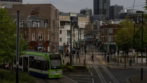 Getty Images A file image showing a wide view of a green and white TfL tram driving on track through Croydon, south London. Roads, overhead cables, trees, traffic signals, buildings and pedestrians can be seen in the background stretching into the distance