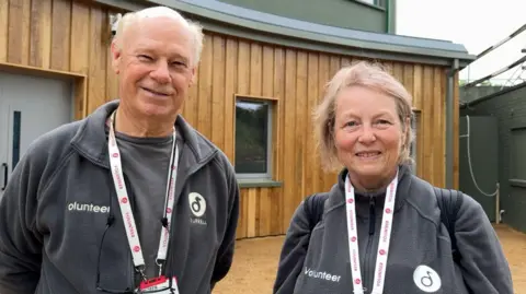 Two people stand side by side outdoors, facing the camera, in front of a building with vertical wooden cladding and green-painted upper sections. Both individuals are wearing grey fleece jackets with the word “Volunteer” embroidered on the front and a circular “Durrell” logo. Each person has a lanyard around their neck with an identification badge attached, and one person is carrying a backpack over one shoulder. Behind them are closed double doors, a small window set into the wooden wall, and a light-coloured gravel pathway. 