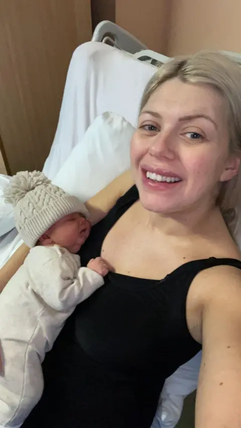 Julie Mclean Julie Scott McLean in a black vest top with blonde hair taking a selfie of herself and a newborn baby in white top and cream woolly hat.