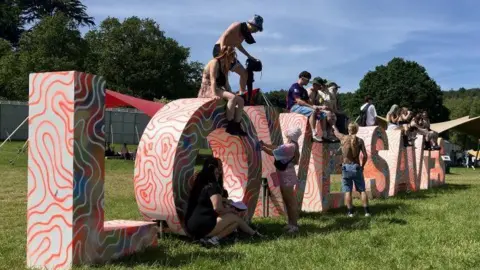 Huge letter blocks decorated in orange squiggly lines spell out Love Saves The Day. The blocks are on green grass and people are climbing over and sitting on them.