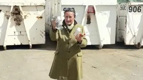 Councillor Elissa Swinglehurst wearing a green tweed coat holds up rubbish in both hands while standing in front of white recycling bins.