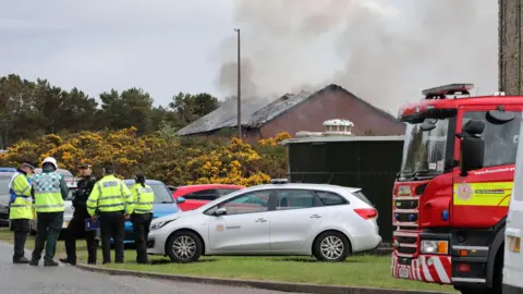 Peter Jolly/Northpix Smoke rises from a hole in the roof of a low, red brick building. The building is obscured by an area of gorse and low circular metal tank. A group of emergency services workers stand in a huddle next to a silver fire service car. There is also a fire engine parked nearby.