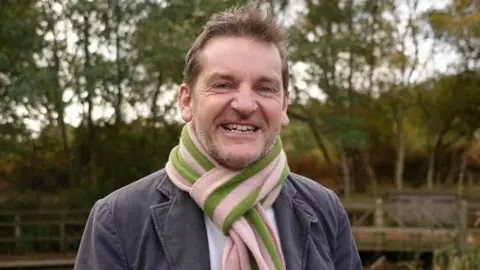 Shaun Whitmore/BBC Pete Waters is wearing a grey coat with lapels over a white shirt which is partially visible beneath a pink and green scarf. He has brown hair and blue eyes and is smiling open-mouthed at the camera with a line of trees behind him.