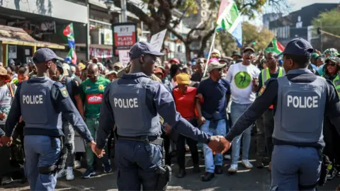 Police officers cordon off an area as protesters chant slogans outside a police station in Pretoria - 30 June 2026.