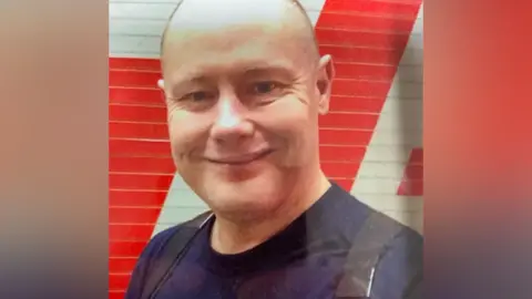 A man is smiling at the camera and has a bald head. He is wearing a navy top and black braces. A metal door behind him is red and white