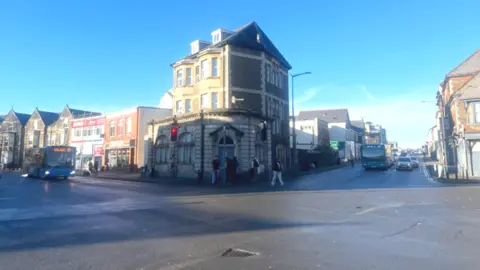 A crossroads, with buildings on each corner, people crossing the road and vehicles on the road. The sky above is blue with bright sunshine, casting shadows on the road.