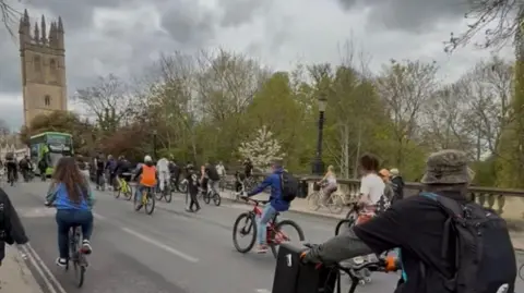 Alex Harding A view of Magdalen Bridge during the bike rave in 2025. Many people are cycling as part of the rave. It's cloudy. A green bus is seen approaching them from the opposite direction.