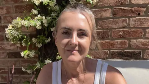 Natalie Heptinstall seated on a cushioned outdoor chair in the foreground, wearing a sleeveless light-coloured top and loose trousers. A necklace and bracelets are visible. Decorative plants, including a red spiky-leaved plant and a climbing plant with white flowers, surround the seating area. A brick wall forms the background.