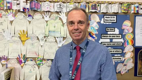 Headteacher Rick Weights stands in front of primary school display board, with paper cut-outs of hooded jumpers that have been coloured in by pupils. Rick has short hair and wears a blue and white checked shirt with a red tie, and a lanyard with the school logo across it.