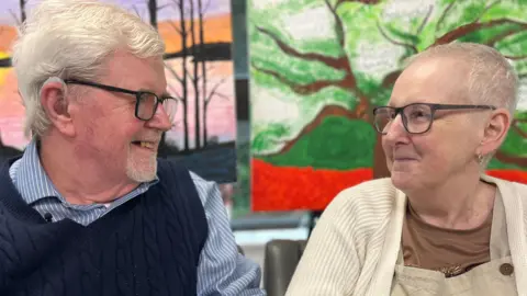 Sally and Kevin are looking at each other and smiling. Kevin is wearing a blue and white striped shirt and a navy knitted tank top. Sally is wearing a cream cardigan, brown t-shirt and light brown dungarees. Both have short white hair and glasses.