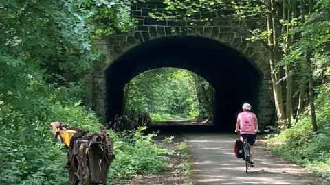 Paul Adams/Walk Wheel Cycle Trust Shot of rural path with a cyclist in a pink top and white helmet riding along it. The path goes under a brick bridge archway. There are trees and hedges all along the sides. 