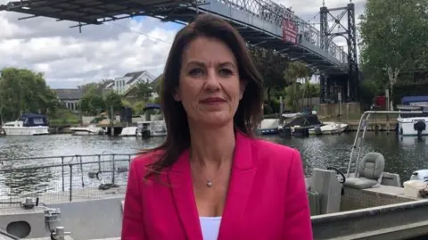 A woman with shoulder length brown hair and a bright pink blazer stands in front of a river. In the background there are boats