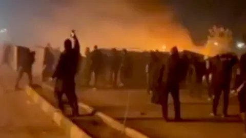 UGC A blurry video grab shows protesters and smoke from a fire, with one person holding up a phone to capture events, taken in Mashhad in early January.