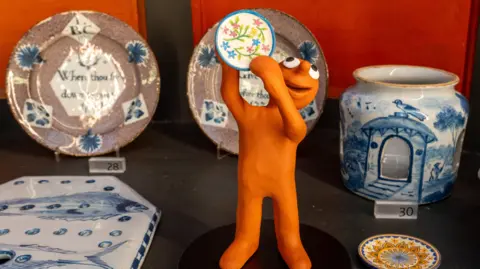 A museum exhibit is filled with plates and white and blue china. Aardman character Morph - a small terracotta-skinned plasticine man - is standing in the middle, holding up a miniature plate of his own.