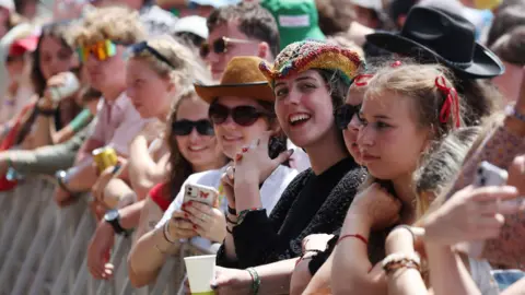 PA Media A row of women in the front of a crowd looking at Glastonbury's main stage. Some of them are wearing hats and sunglasses.
