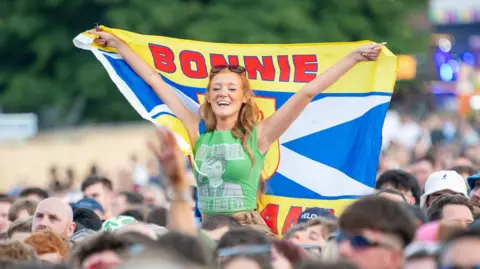 A music festival crowd. A woman with red hair and sunglasses on her head sits on another reveller's shoulders, holding a flag. It has the Scotland flag and lion rampant on it, with BONNIE SCOTLAND printed in large letters 