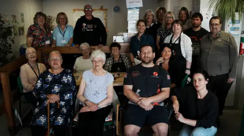 John Fairhall/BBC A group photo of men and women all of different ages. Some are sitting and some are standing. They are all smiling and looking into the camera. 