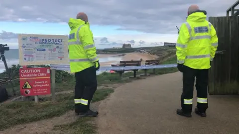 BBC Coastguard members stand behind police tape at Fistral