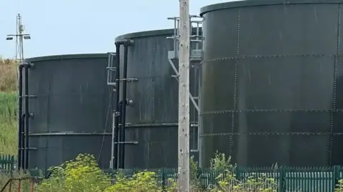 Three large metal black cylindrical waste tanks behind a green metal fence