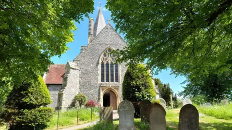 The exterior of St Andrew's Church in Alfriston, a 14th century building with a steeple and several graves in the foreground.