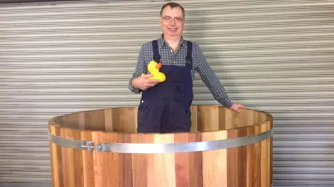 Alan Paulus Alan Paulus wearing building wear and standing in a large wooden hot tub, while holding a rubber duck.