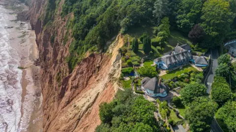 James Loveridge/BNPS A large house with a manicured garden sits close to the edge above the sea with more houses further inland and nearby hedges and trees and a road.