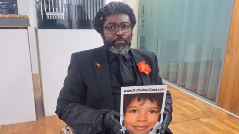 Nadia Lincoln/BBC Kye Gbangbola sitting in a wheelchair. He is wearing a black suit, shirt and tie and holding a poster of Zane.