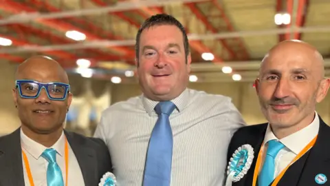 Phil Shepka/BBC Karan Maheshwari, Ryan Coogan and Howard Tobias standing in a sports hall. Karan is wearing a grey suit, with a blue and white Reform rosette, a white shirt, a bright blue tie and blue framed glasses. Ryan has short dark brown hair and is wearing a striped shirt and blue tie. Howard is wearing a black suit, with a blue and white Reform rosette, over a white shirt and bright blue tie. 