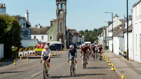 A group of cyclists pedal through the heart of the south of Scotland town of Gatehouse with houses down both sides of the road and traffic cones and a tower with a clock on it in the background.