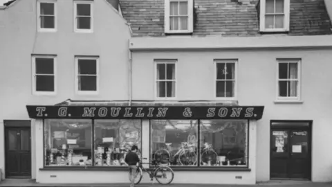 Chris George A black and white wide shot of the bicycle shop, T. G. Moullin and Sons. It has a large sign with the shop's name across it, and the front window is filled with bicycles.