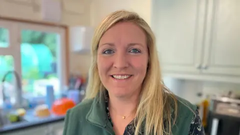 Laura Foster/BBC Milly Fyfe stands in a kitchen. She has blonde hair and is smiling into the camera. She is wearing a green fleece zip-up vest over a black patterned blouse.