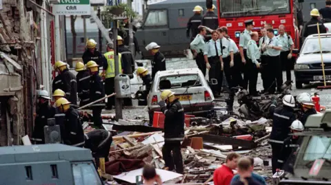 Firefighters and police officers attending to the aftermath of the Omagh Bomb. There is a white car amongst the wooden and metal rubble, with a fire engine, another black car and van in the background.