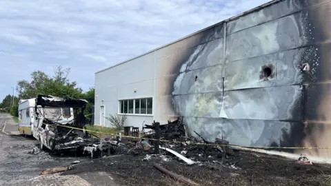 BBC The burnt-out shell of a caravan can be seen parked next to a pile of charred debris, which appears to be another caravan completely destroyed by the fire. The cladding on the side of the store is heavily burnt, with the metal panels stripped of paint and contorted by the fire. 