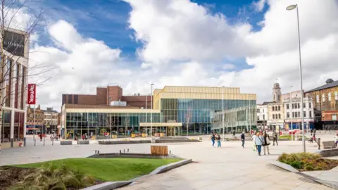 Barnsley Council The image shows a modern town square with a spacious, open design featuring light-coloured paving stones and landscaped areas with grass, plants, and small shrubs. There are a few wooden benches placed around the square, providing seating for visitors. Several people are walking across the square, some carrying shopping bags.
In the background, there is a large contemporary building with tall glass windows and a golden-beige facade, which appears to be a central feature of the square. To the left, a TK Maxx store is visible with its red and white signage. Surrounding the square are older, traditional buildings with brick and stone exteriors. The sky is partly cloudy with patches of blue.