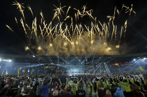Getty Images Fireworks light up the sky during the closing ceremony of the 2014 Commonwealth Games at Hampden Park. A large crowd is gathered outside the stadium and many people are raising their hands in celebration.