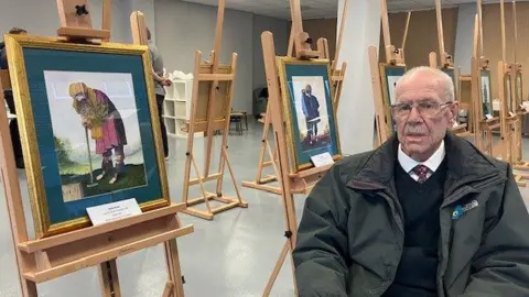 Dick Scott, with balding grey hair and a moustache, wearing glasses, a green coat, navy jumper, white shirt and tie, sits in a chair next to a series of watercolour paintings on a wooden easels. 