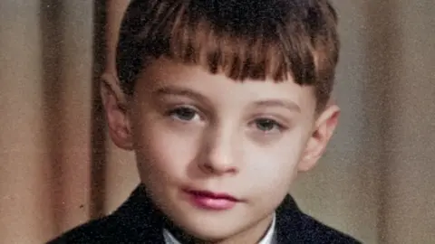 A young man with brown hair with a straight fringe looking straight into the camera