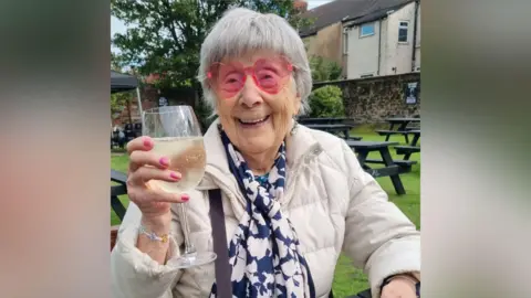Anne has short grey hair and is wearing pink heart shaped sunglasses and holding a glass of white wine. She is sat in a beer garden and smiling at the camera.
