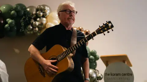 Karol McGonigle Paul brady holding a guitar. He is wearing a black t-shirt and glasses and has white hair. There is a balloon arch on the left and a podium on the right with a sign saying 'the koram centre GRAND OPENING'